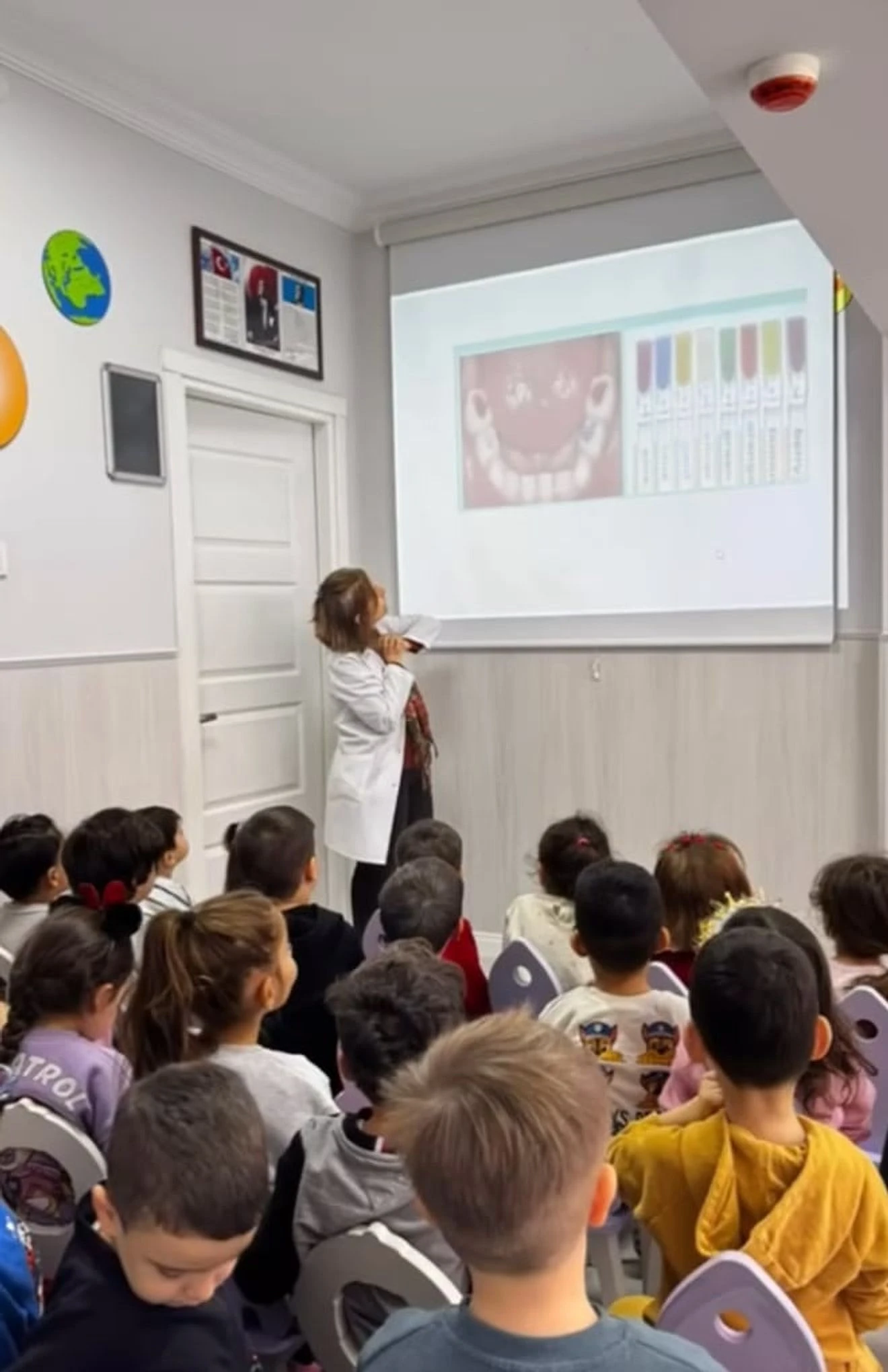 Beyaz önlüklü uzmanın diş sağlığı sunumunu izleyen çocuklar